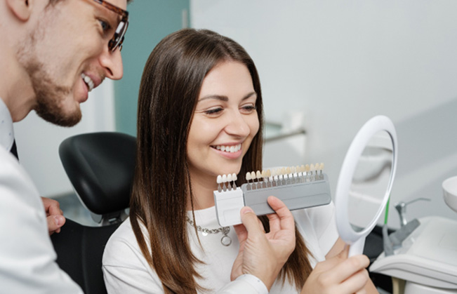 Dentist holding shade guide to woman’s smile as she looks at reflection in mirror